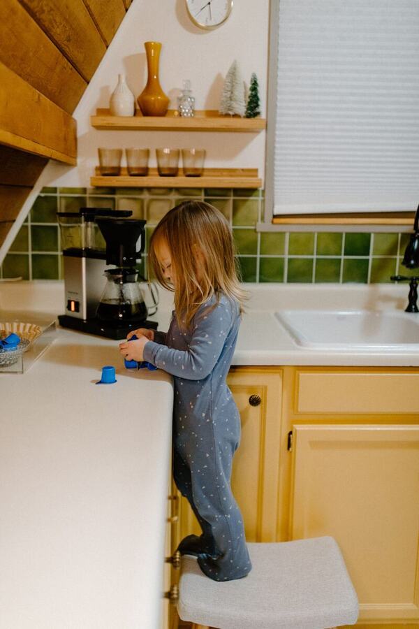 A child helping at the kitchen counter on a step stool beside the green tile backsplash