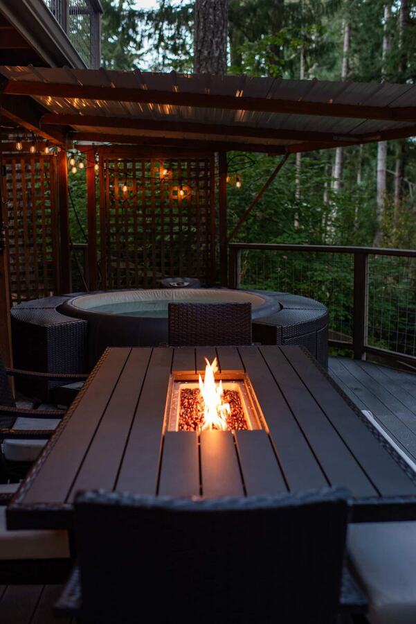 The covered deck at night with the hot tub and the long fire pit dining table